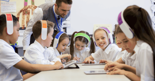 A group of children at primary school wearing headphones smile as they use tablets while their teacher watches.