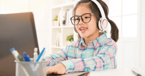 Kleines Mädchen mit Kopfhörern beim Sitzen auf einem Schreibtisch auf einem Laptop