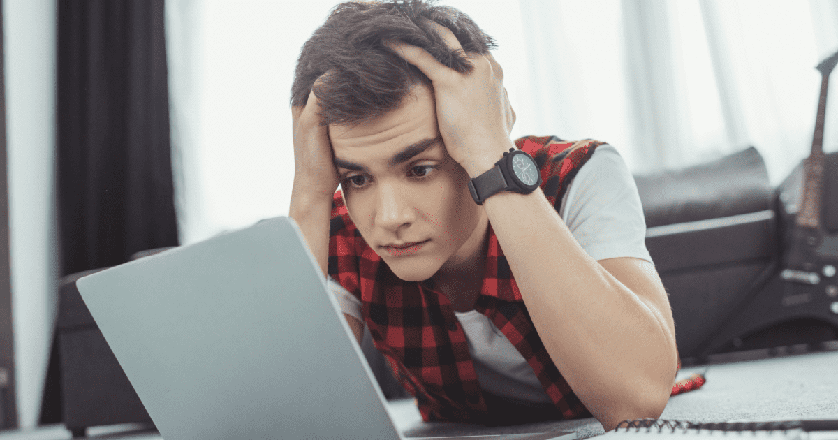 Teen boy holds his head and looks concerned as he looks at his laptop.