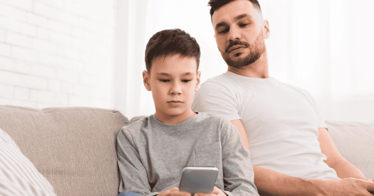 A father watches over a son's shoulder as he browses his smartphone, both with neutral expressions
