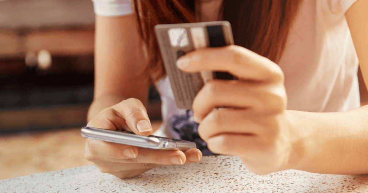 一个年轻女孩一只手拿着智能手机，好像在浏览，另一只手拿着信用卡，可能成为网络诈骗的受害者