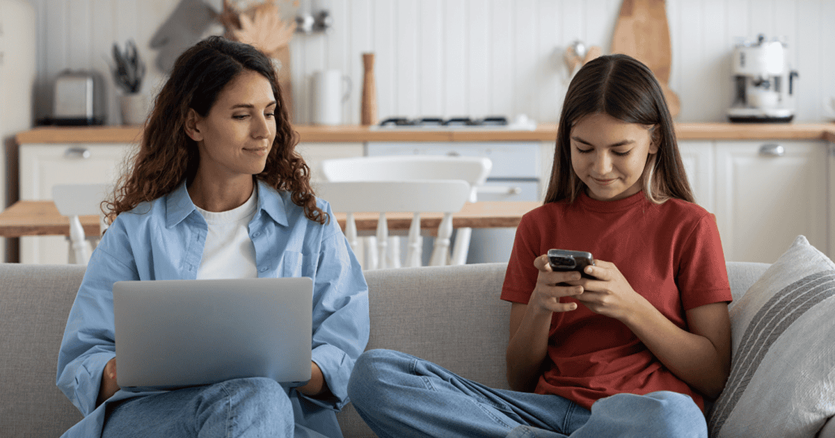 Mãe e filha sentadas no sofá, a mãe com um laptop e sorrindo, olhando para a filha enquanto a filha sorri para o smartphone nas mãos.