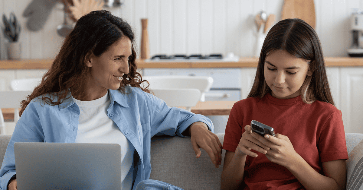 Mutter mit Laptop lächelt ihre Tochter im Gespräch an, während sie ihr Smartphone durchsucht.