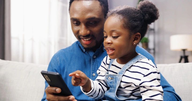 A father looks at a smartphone with his daughter, an important part of digital safety in addition to parental controls.