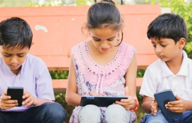 Zdjęcie pokazuje troje dzieci siedzących na zewnątrz i korzystających z urządzeń. Wybór odpowiednich aplikacji może wspierać dobre samopoczucie i zrównoważony czas przed ekranem.