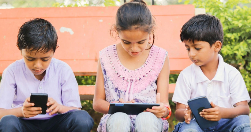Uma foto mostra três crianças sentadas do lado de fora e usando dispositivos. Escolher os aplicativos certos pode ajudar no bem-estar e no tempo de tela equilibrado.