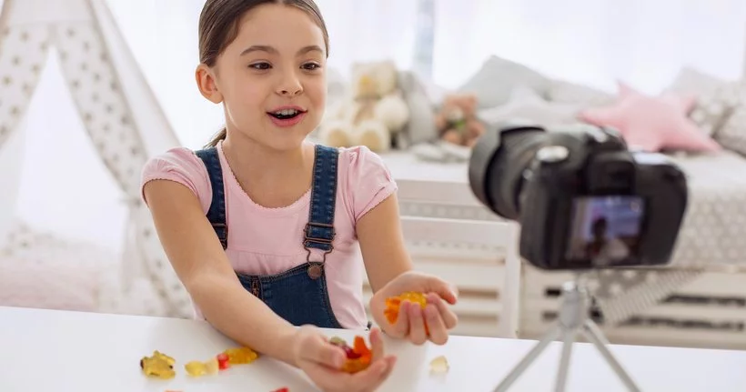 Маленький ребенок с камерой на штативе ведет прямую трансляцию или ведет видеоблог со сладостями.