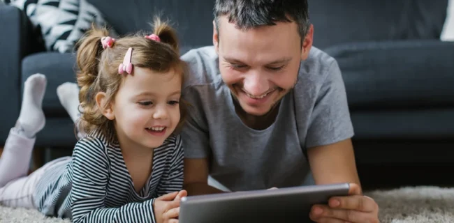 A dad and toddler play on a tablet, laughing.
