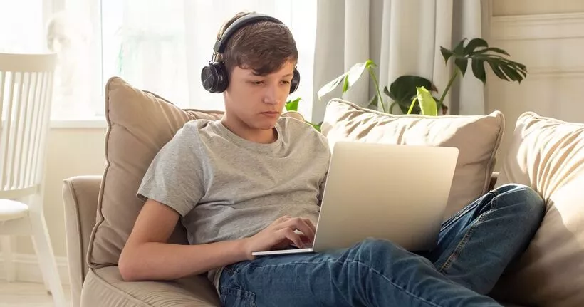 A teen boy uses a laptop while sitting on the sofa.