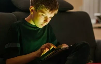 एक लड़का अपना स्क्रीन समय टैबलेट पर बिताता है और उसके चेहरे पर प्रकाश प्रतिबिंबित होता है।