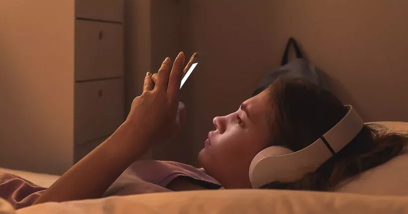 Подросток носит наушники, лежит и смотрит в телефон.