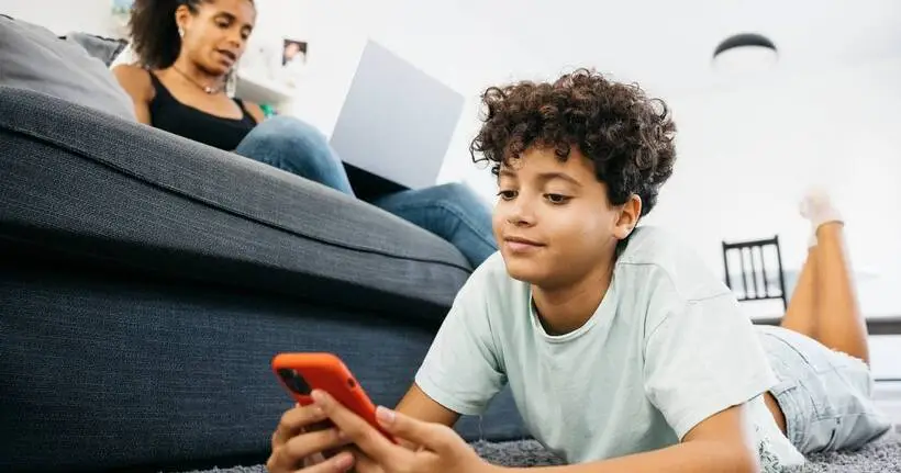 A boy uses his phone on the floor while his mum uses her laptop on the sofa.
