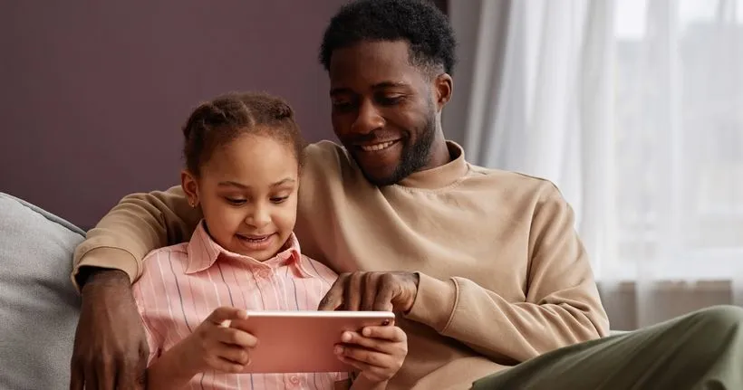 A dad sits with his daughter while she uses a device.