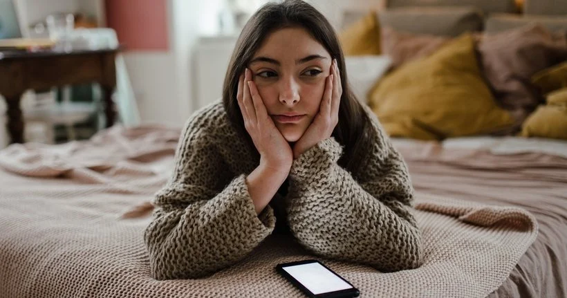 一个女孩躺在床上，她的智能手机放在她面前，她可能很沮丧。