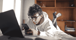 A teen browses his smartphone in front of a laptop.