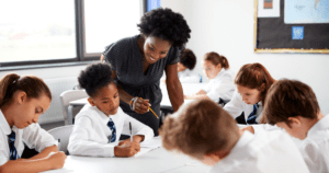 A female teacher smiles and helps a group of students working at their table.