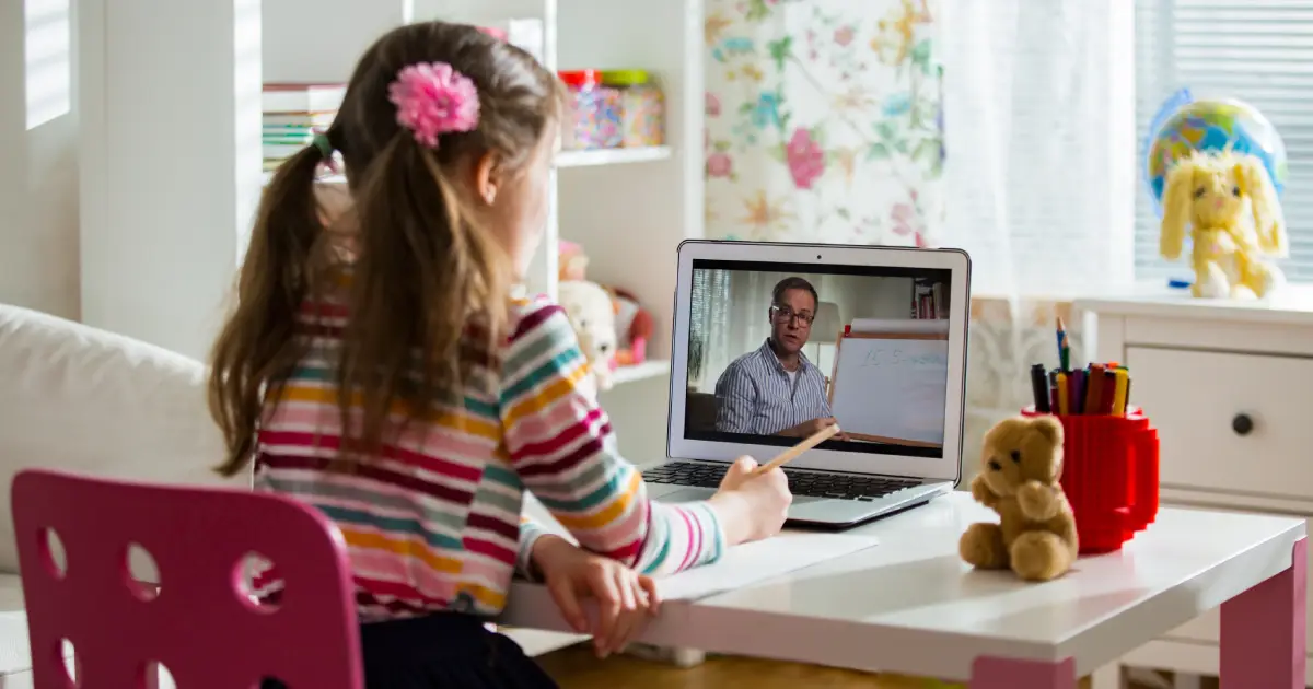 A young child sitting at a desk watching an online lesson on a laptop, holding a pencil while a teacher appears on the screen.