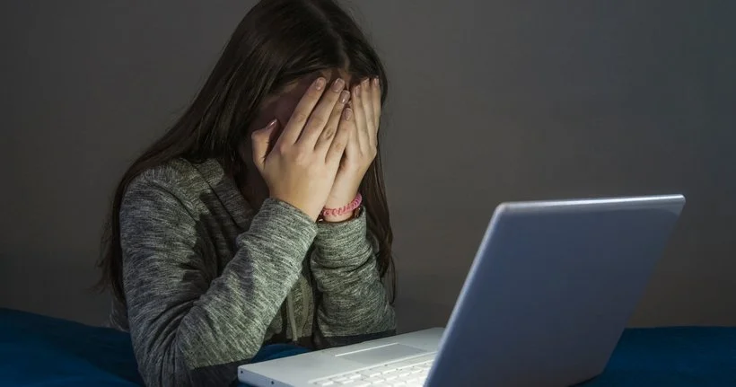 A girl sits in front of a laptop in the dark with her face covered by her hands.
