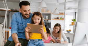 An adult and a child using a tablet at a table, with another adult and a child studying in the background.