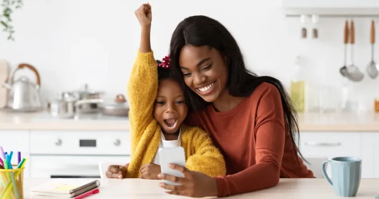 A woman holds a smartphone while having a child sitting on her lap. Both are smiling and looking at the device's screen