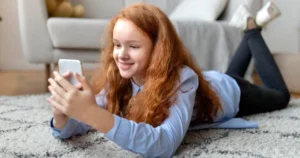 Une jeune fille est allongée sur le ventre et consulte son smartphone.