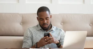 A dad browses his smartphone with his laptop open next to him.
