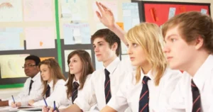Six students sit in a classroom with one raising their hand during a lesson.