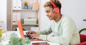 A teen boy uses his laptop while wearing headphones. His smartphone sits nearby.