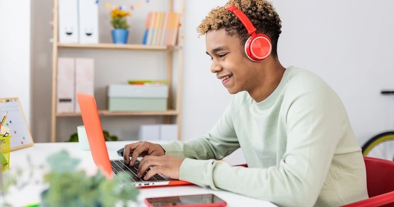 A teen boy uses his laptop while wearing headphones. His smartphone sits nearby.
