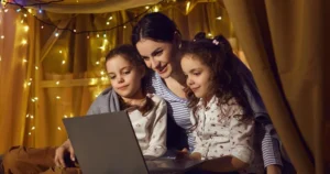 A mother looks at a laptop with two daughters inside a homemade blanket fort.