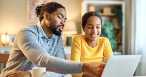 A dad instructs his daughter as they look at a shared laptop.