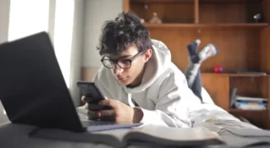 A boy lying on his bed using his phone, while his laptop sits open in front of him