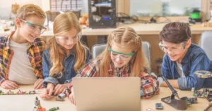 Four children gather around a laptop with pieces of circuit boards and similar around them while they wear protective glasses.