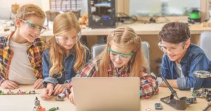 Four children gather around a laptop with pieces of circuit boards and similar around them while they wear protective glasses.