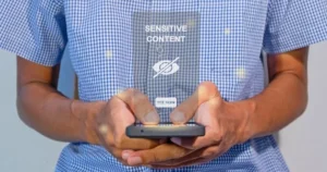 Someone holding a smartphone with a graphic hovering overtop which reads 'sensitive content'.