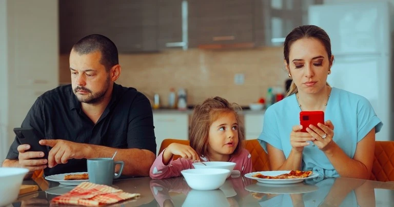 A mother and father use their phones at the dinner table while their child watches.