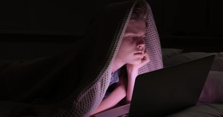 A boy sits under a blanket on his stomach, looking concerned at a laptop.