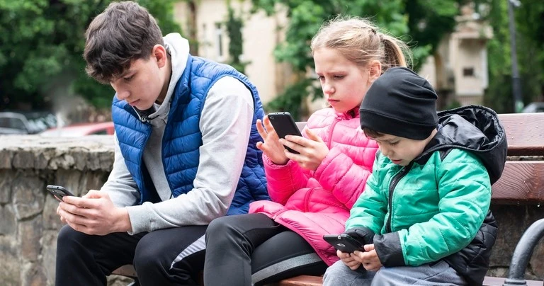 Three different aged children use smartphones outside.