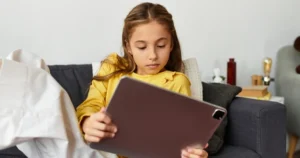 A young girl lies on a sofa watching something on her tablet.